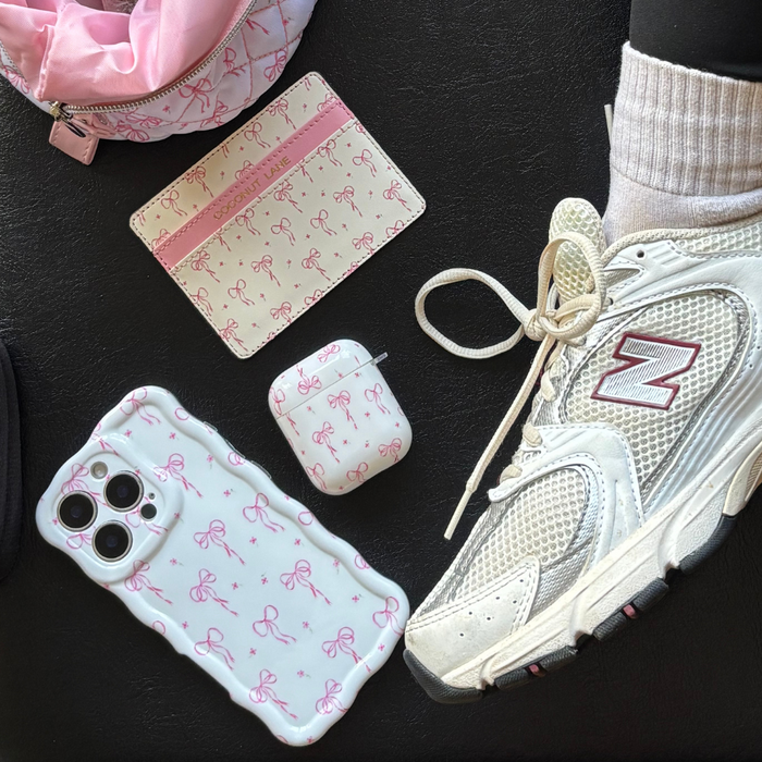 Coconut Lane AirPods case with pink dainty bows, white New Balance sneaker, and iPhone on a black surface.