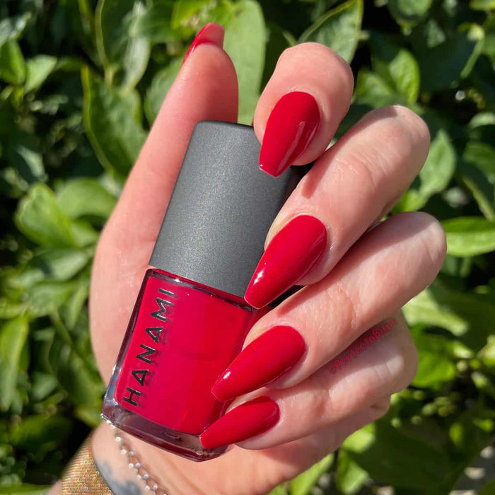Hand holding a red nail polish bottle with green foliage in the background.