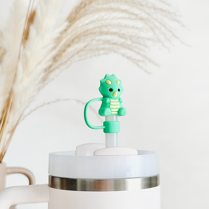 Green dinosaur straw cover on a white tumbler, background shows softly blurred beige pampas grass.