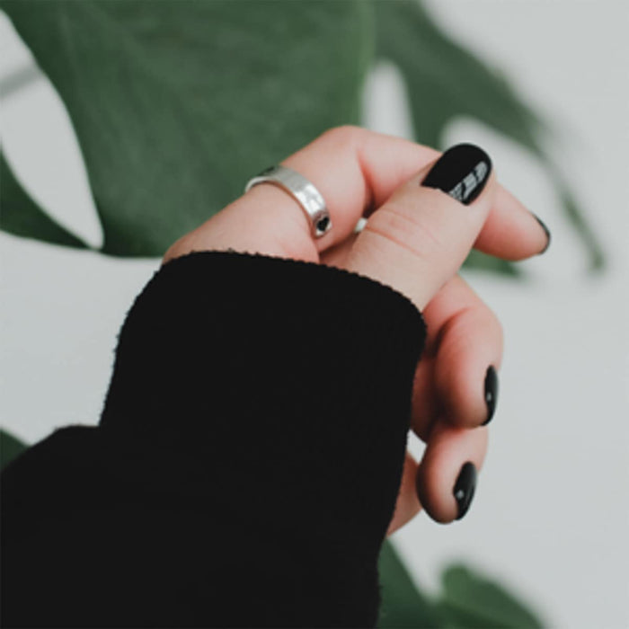 Hand with black nail polish and silver ring against a blurred green leaf background.