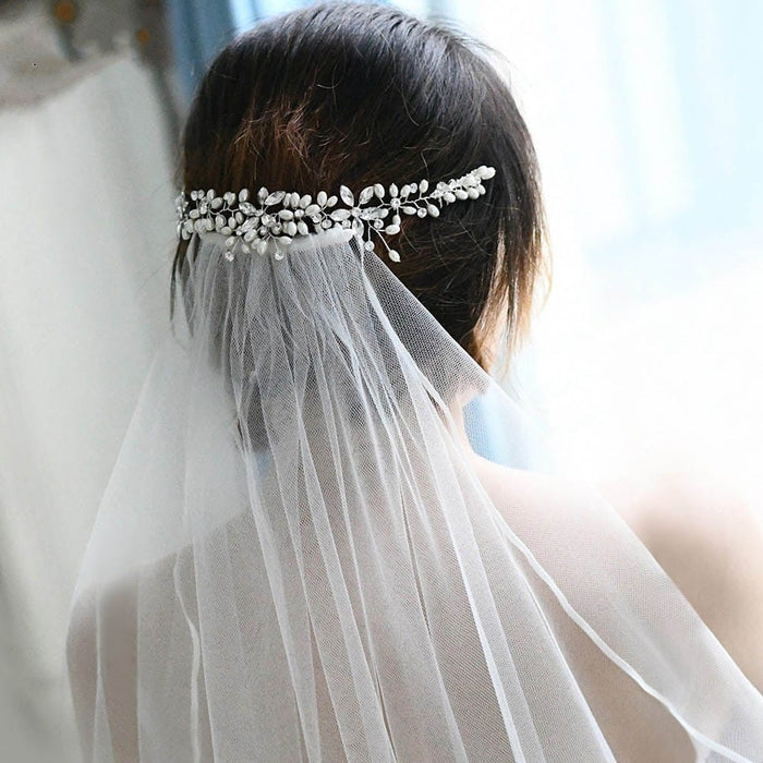 A person with short brown hair wears a white veil, secured by a decorative silver hairpiece with pearl and crystal accents, seen from behind in soft natural light.