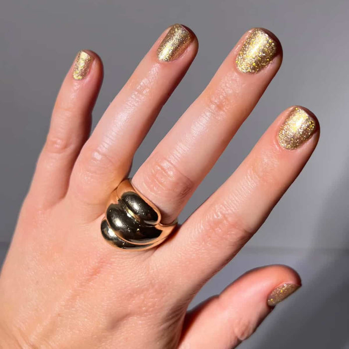 Hand with gold nail polish and a gold ring on a neutral background.