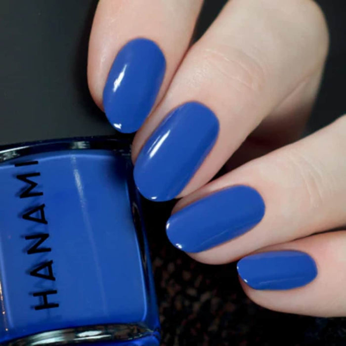 Hand with blue nail polish holding a blue nail polish bottle labeled 'HANAMI'.