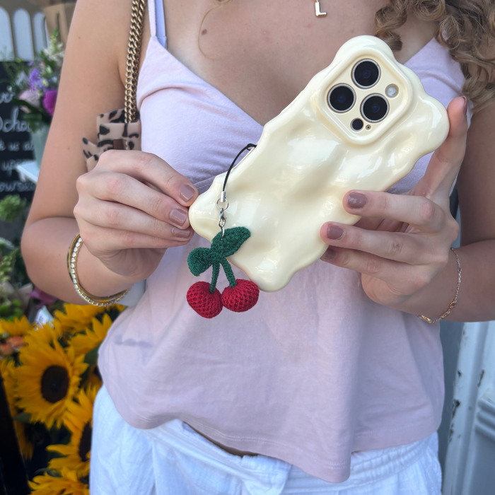 Woman holding wavy cream phone case with red cherry charm; sunflowers in the blurry background.
