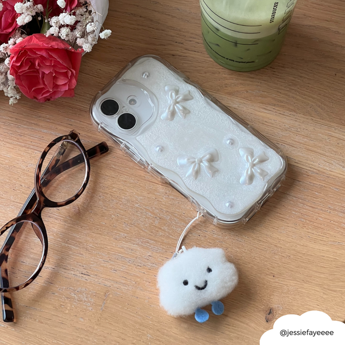 Phone with a white bow case and cloud charm on desk next to glasses, flowers, and a green drink.