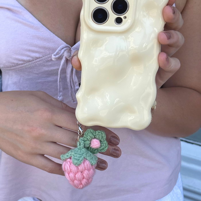 Hand holding a phone with a wavy cream case and a pink strawberry phone charm.
