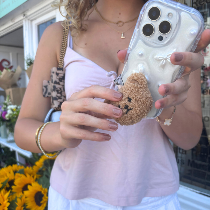 Woman in a light pink top holding a phone with Coconut Lanes teddy bear phone charm; sunflowers in the background.