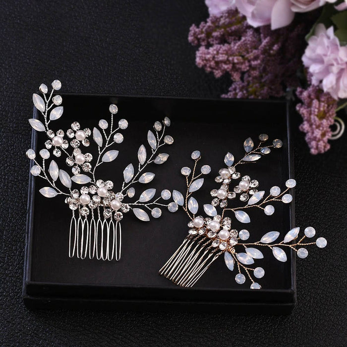 Two elegant silver hair combs with pearl-like beads and crystal leaves in a black box, with purple and pink flowers behind.