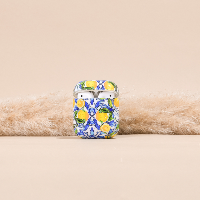 The AirPods Case - Amalfi Lemon by Coconut Lane, crafted from premium materials, showcases a vibrant floral and lemon pattern. It rests on a soft, white, fluffy surface against a neutral beige background. The top of the case is slightly open, revealing the white AirPods inside.