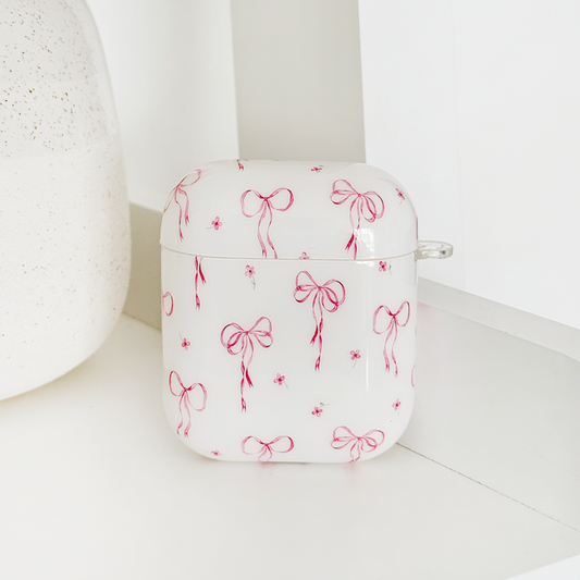 Coconut Lane AirPods case with a dainty bows pattern on a white shelf next to a vase.
