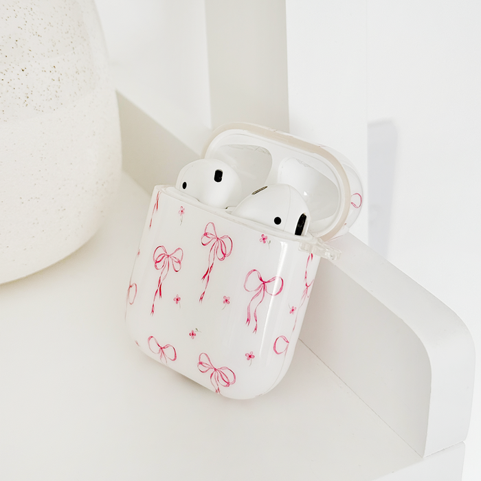 White earbuds in a Coconut Lane AirPods case with pink bow pattern, resting on a white shelf.