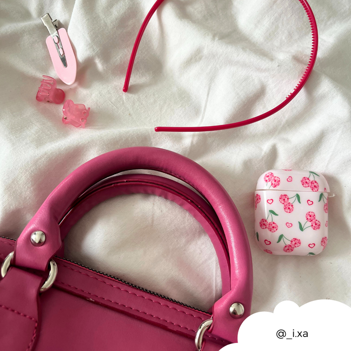 Pink headband, assorted hair clips, cherry print AirPods case, and pink handbag on white fabric background.