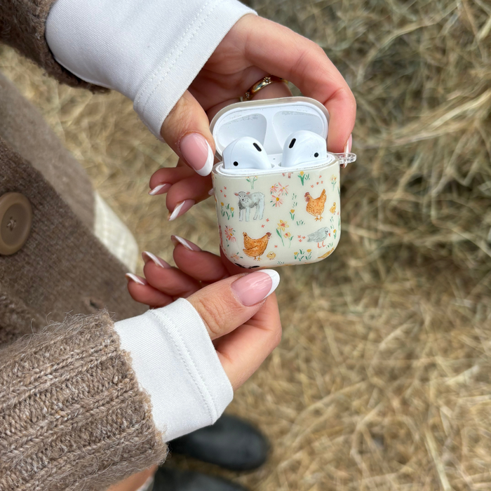 Hands hold a Coconut Lane Airpods case with a farmyard pattern of chickens and flowers on a cream background.