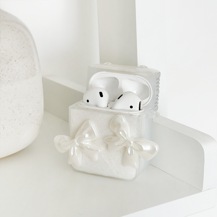 White wireless earbuds in a pearly Coconut Lane Airpods case with bows, resting on a white shelf next to a round object.