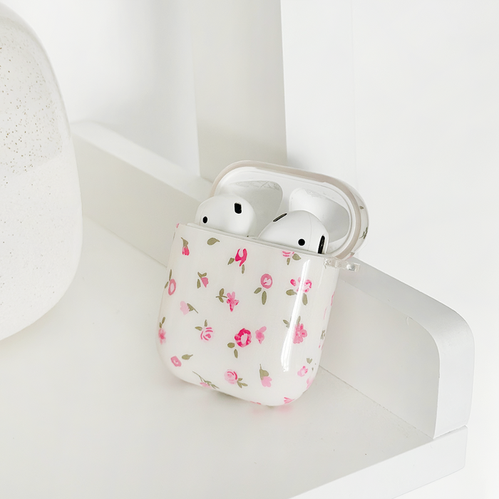 White wireless earbuds in a pink floral Coconut Lane AirPods case, resting on a white shelf next to a round object.