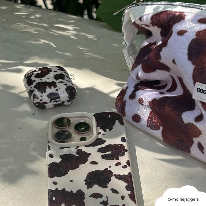 Brown and white cow print AirPods case, phone, and bag on a sunlit surface with green leaves.