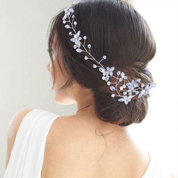 A woman with dark hair in a low bun wears a delicate silver and pearl floral headpiece, shown from behind in a white off-shoulder bridal gown.