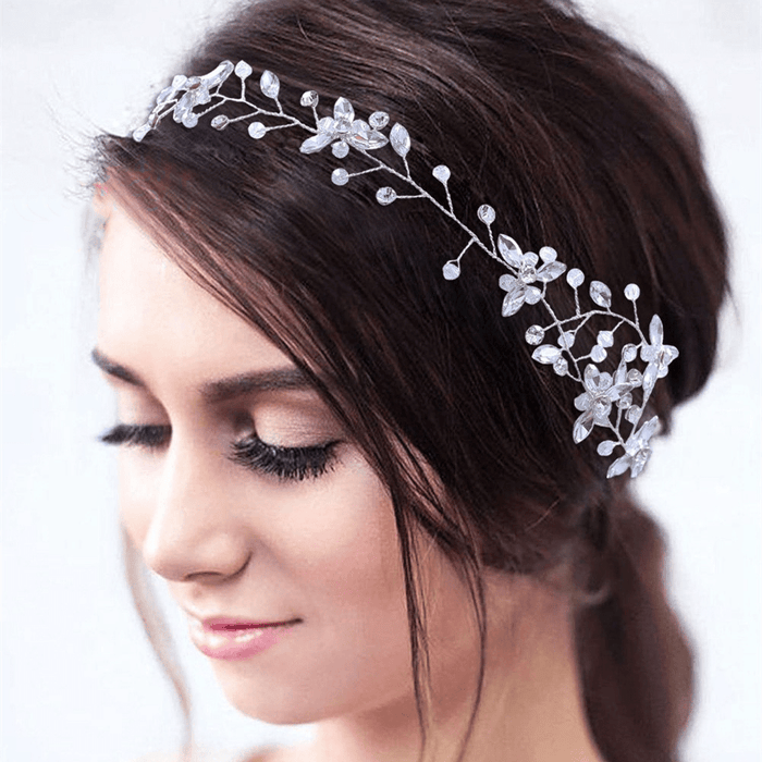 A woman with long dark hair styled up wears a delicate silver floral headband with crystals and pearls, smiling gently, dreamy background.