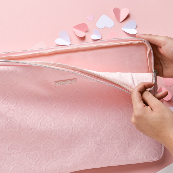 Hands unzip a baby pink heart-patterned laptop sleeve on a pastel pink surface with paper heart cutouts.