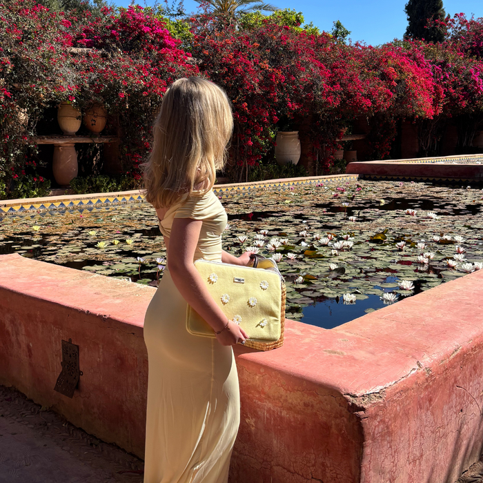 Woman in a light yellow dress holds a floral Coconut Lane Buttercup Bloom laptop sleeve by a lily pond with pink flowers.