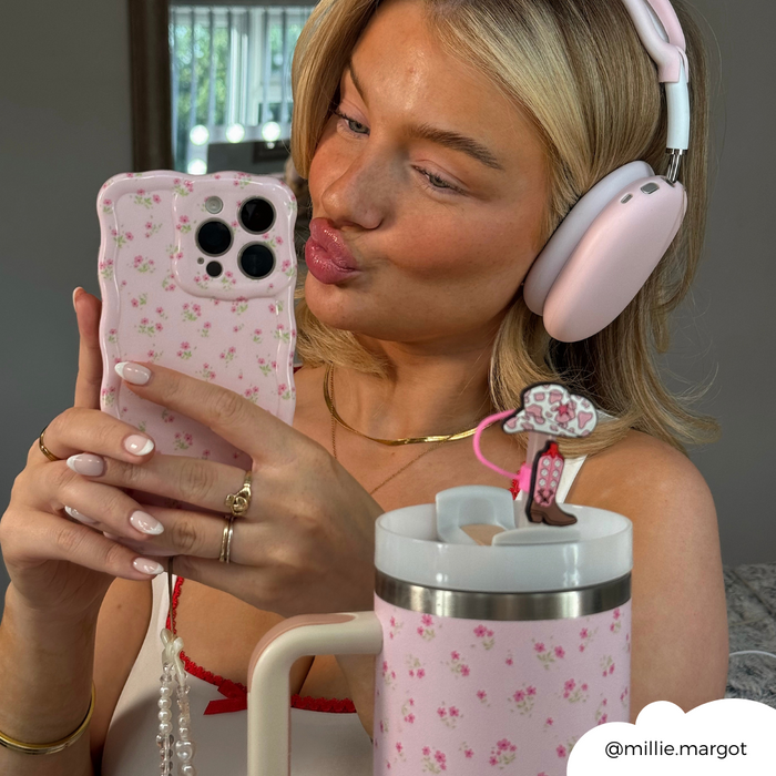 Blonde woman in pink headphones takes a mirror selfie with a floral phone case and straw cup cover featuring western accents.