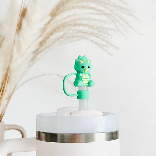 Green dinosaur straw cover on a white tumbler, background shows softly blurred beige pampas grass.