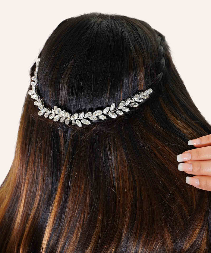 A woman with long, dark hair in loose waves wears a sparkling silver leaf-shaped hair accessory. Her manicured nails gently touch her hair.