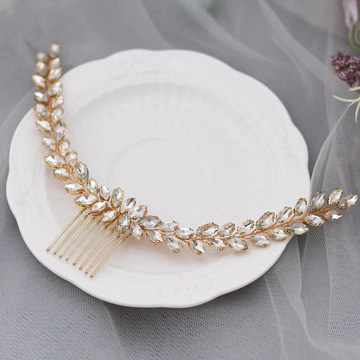 A gold hair comb with clear, marquise-shaped crystals in a leafy pattern rests on a white plate, sheer gray fabric behind.