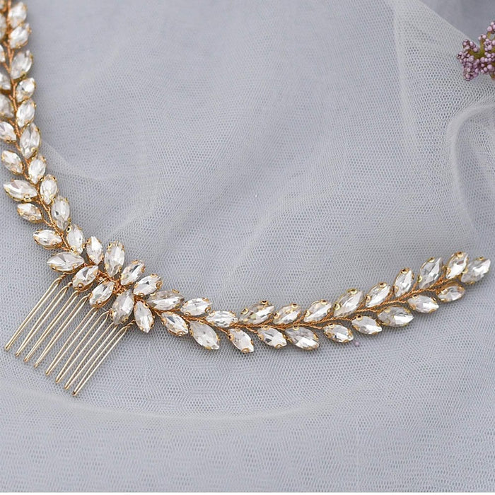 A gold hair comb adorned with leaf-shaped clear rhinestones rests on gray tulle, with a small cluster of purple flowers nearby.