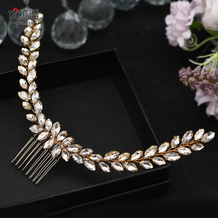 A gold hair comb with two curved rows of marquise-shaped clear crystals arranged like leaves, on a black surface with flowers and glass beads.