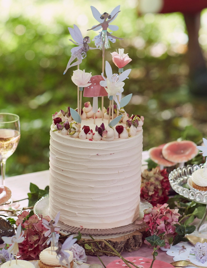 Decorative cake with fairy toppers on a table with flowers and a glass of wine.