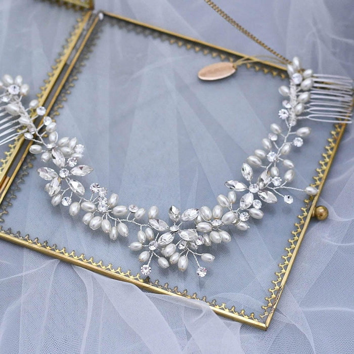 A delicate bridal hair accessory with pearl and crystal floral designs rests on a gold-edged glass frame atop soft white tulle fabric.