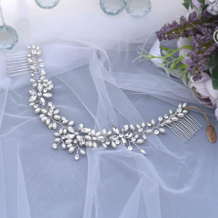 A delicate silver bridal hairpiece with pearls and crystals in a floral design, on white tulle beside a bouquet and candle holders.