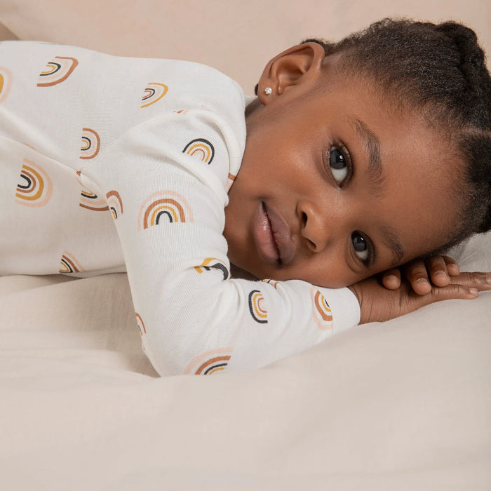 Girl lounging in Petit Lem Off-White rainbow pajamas with elastic cuffs and comfortable full-length pants.