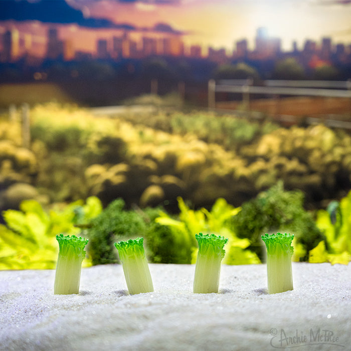 Four glow-in-the-dark itty bitty celery stalks stand in a sandy foreground with a blurred cityscape and greenery in the background, their delicate green contrasting beautifully against the urban backdrop.