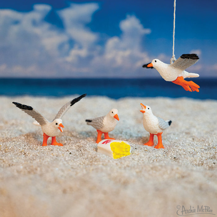 The Itty Bitty Flock of Seagulls features four seagull figurines on sand around a fry container, with one soft vinyl seagull hanging by a string above.