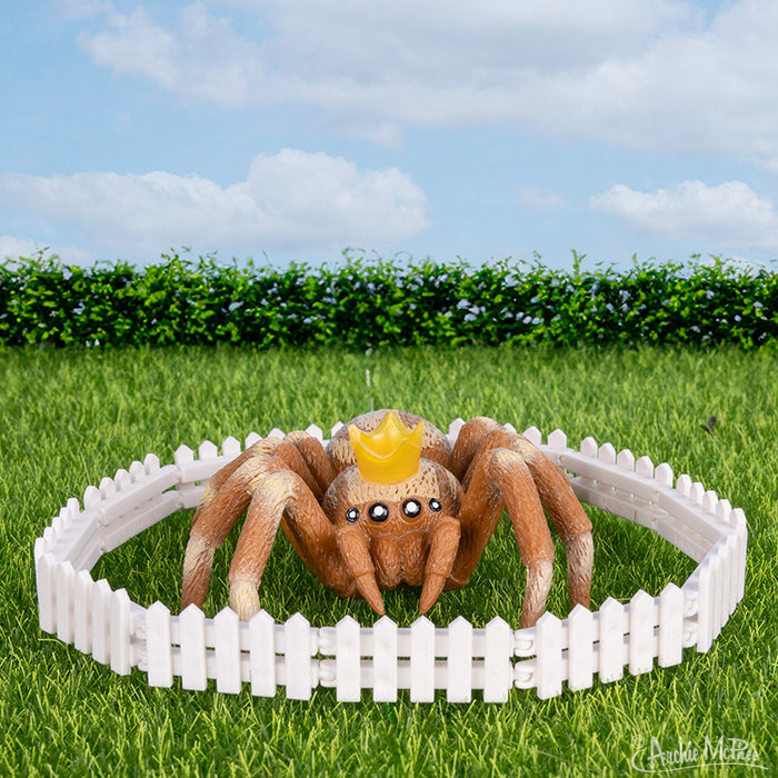 A toy spider wearing a crown rests on the grass, encircled by an Itty Bitty White Picket Fence. In the background, a lush hedge and clear blue sky create a charming scene. Made from soft vinyl, this delightful setting sparks the imagination.