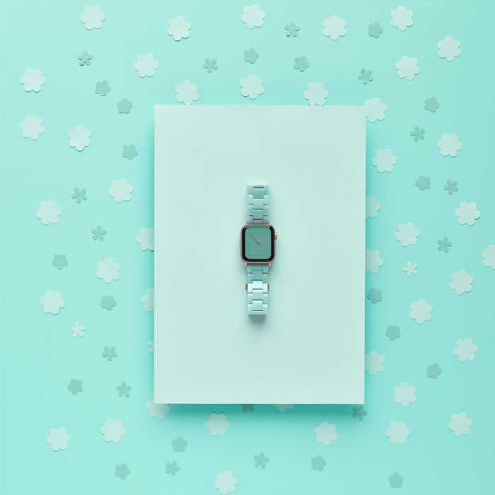 A minimalist photo of a light blue smartwatch featuring the Luxe Pastel Mint Apple Watch Strap by Coconut Lane, centered on a light blue rectangular surface. The background is a similar shade of blue, adorned with numerous small, white, cloud-like shapes scattered around.