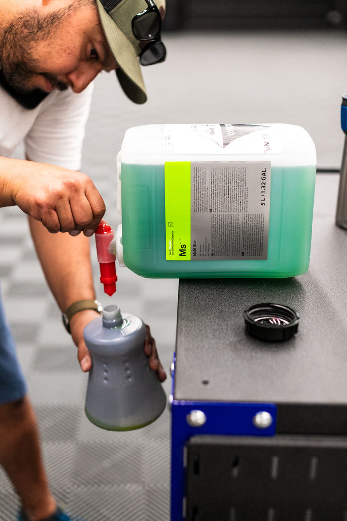 Person pouring liquid from a large container of KochChemie Multi Star into a iK Sprayer bottle on a workbench.