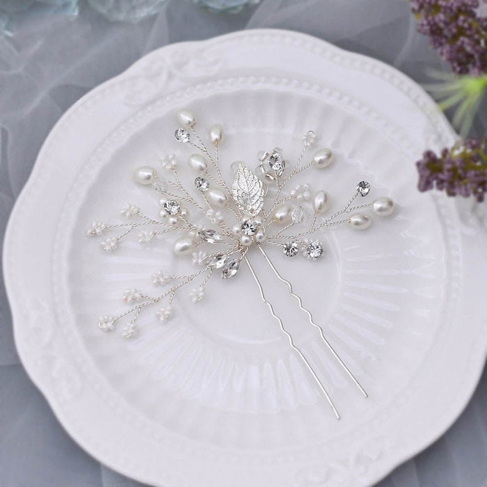 A delicate silver hairpin with pearl and crystal accents rests on a white ornate plate, surrounded by soft lavender flowers and sheer fabric.