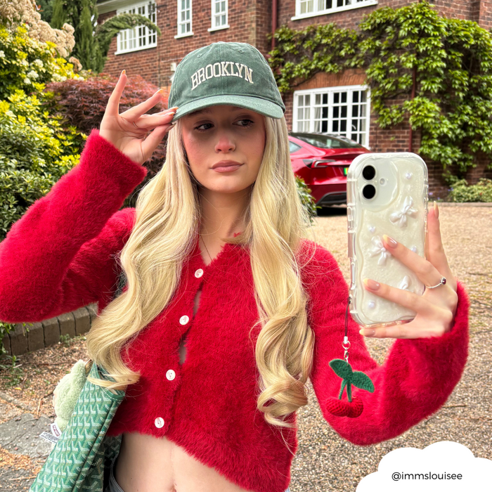 Blonde woman takes a selfie outdoors, phone with red cherry beaded phone charm and wearing a black Brooklyn cap.