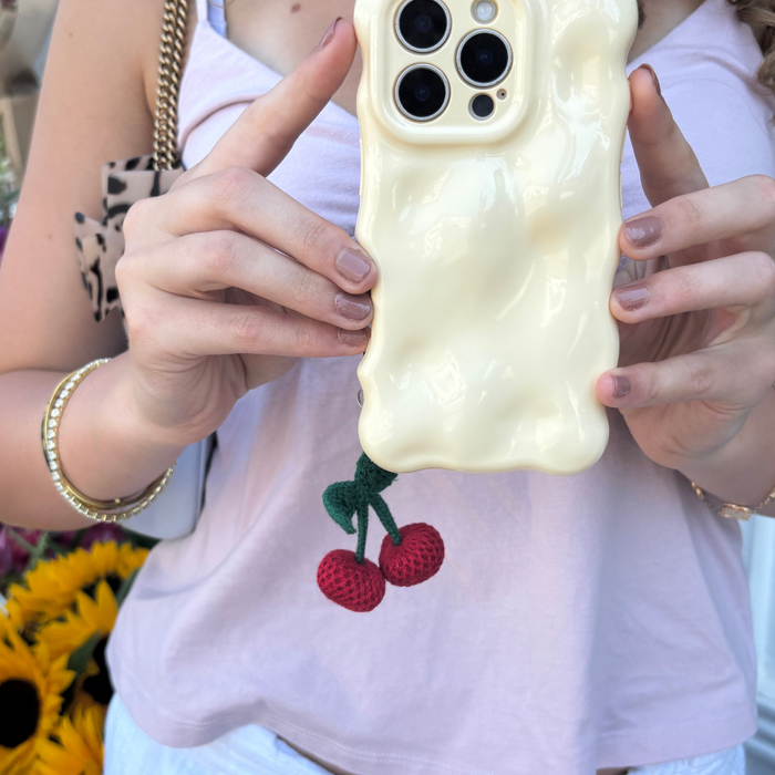 Woman holds a phone with a wavy cream case and Coconut Lane Cherries Phone Charm, wearing a tank top and gold jewelry.