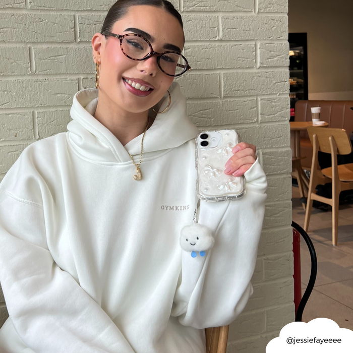 Woman in glasses and white hoodie holds a phone with a pastel Cloud Phone Charm at a cozy cafe.