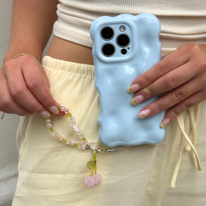 Hand holding a phone with a blue textured case and pink cherry charm, person wearing light yellow shorts.
