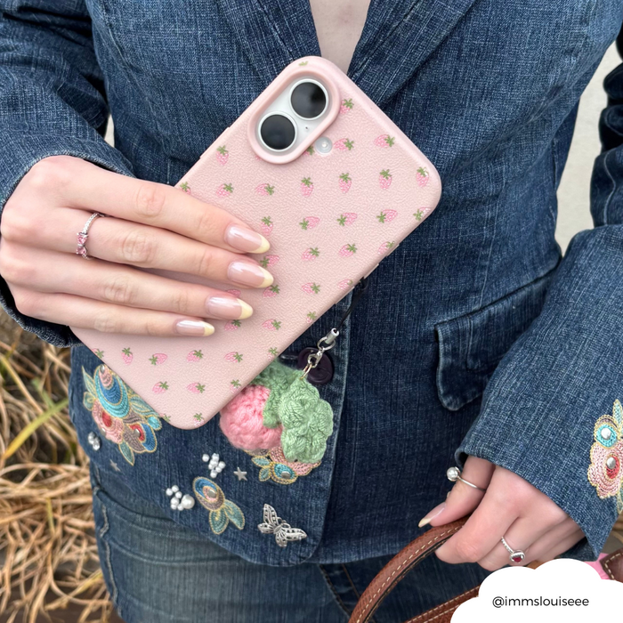 Pink phone with Coconut Lane Pink Strawberry phone charm held by person in denim jacket, manicured nails visible.