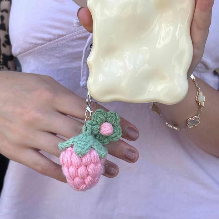 Hand holding a cream phone case with a pink crocheted strawberry charm attached.