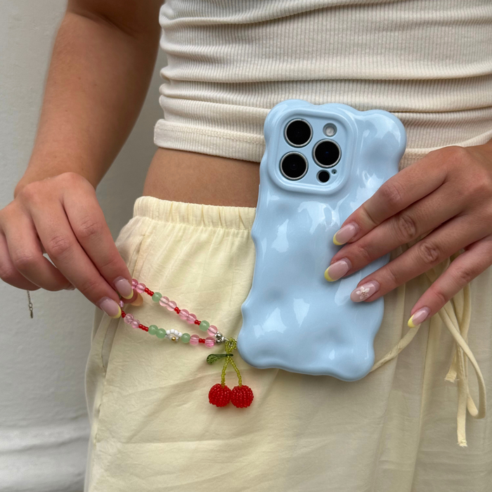 Blue wavy phone case with red cherry charm, held by a person in cream pants and a white top.