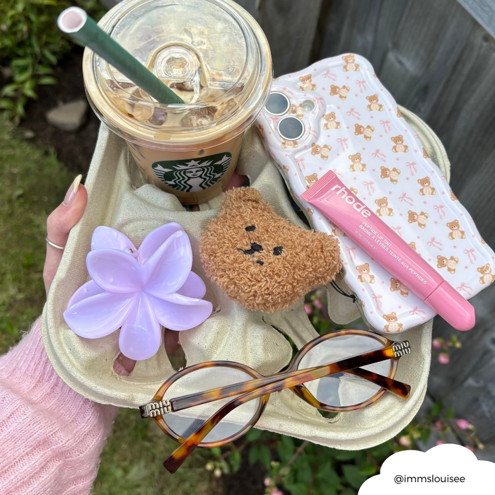 Hand holds tray with iced coffee, sunglasses, flower hair clip, lip balm, and a beaded teddy bear phone charm.