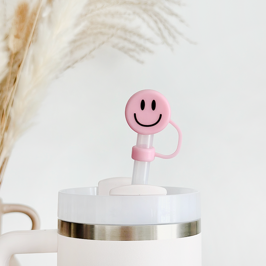 Pink smiley face straw cover by Coconut Lane on a straw in a white cup, with pampas grass blurred in the background.