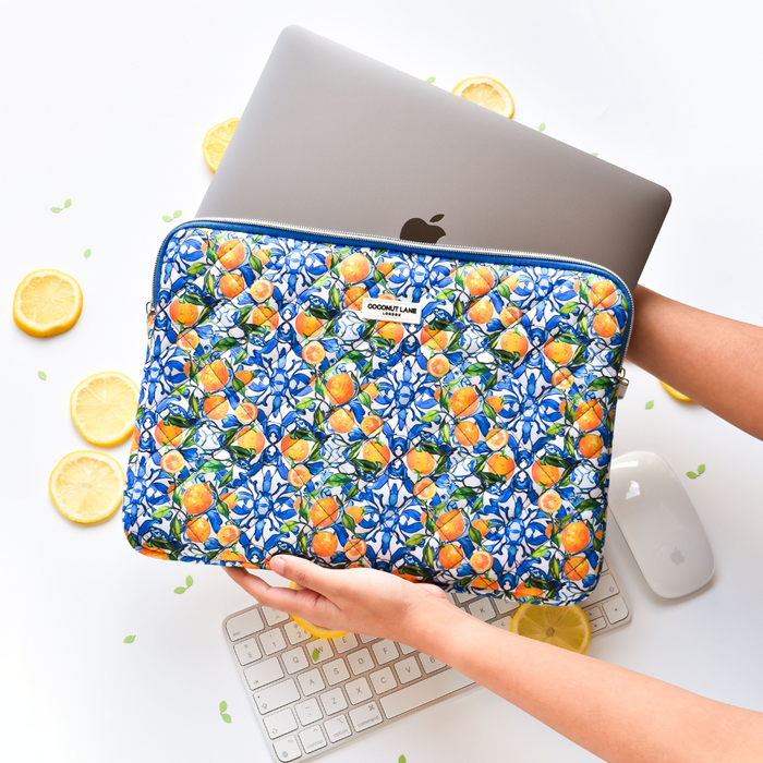 Hands holding a yellow quilted laptop sleeve with lemon pattern above a keyboard, mouse, and lemon slices.